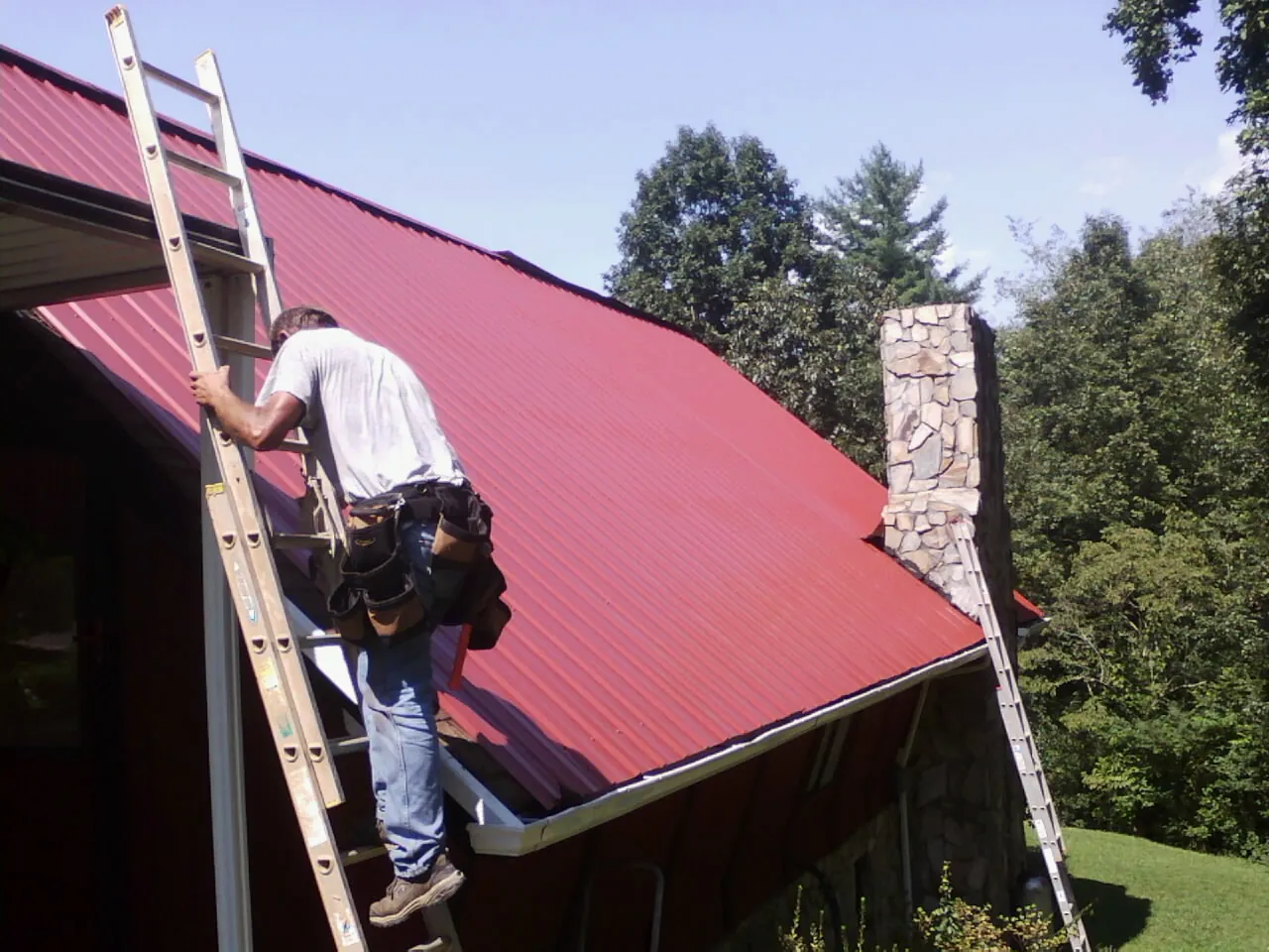 Roofer on a ladder beside a red metal roof and stone chimney — Soffit & Fascia Repair by Roofing Brothers in Beltsville